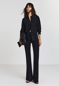 Donna con capelli lunghi che indossa una camicia nera di pantaloni larghi, pantaloni neri svasati, tacchi open-toe, che tiene una pochette nera contro uno sfondo semplice.