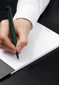 Zelená tužka připravená nad prázdným bílým papírem, ležící na černém texturovaném povrchu, s černou koženou složkou částečně viditelnou na okraji.