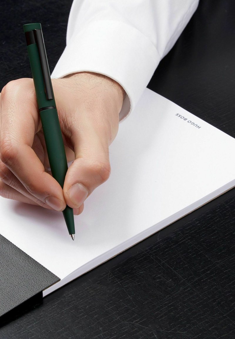 Zelená tužka připravená nad prázdným bílým papírem, ležící na černém texturovaném povrchu, s černou koženou složkou částečně viditelnou na okraji.