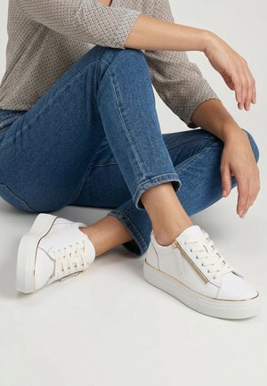 Person siddende med benene over kors iført blå jeans, mønstret beige langærmet skjorte og hvide platformsneakers med lynlås og snører på siden.
