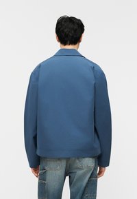 Άντρας που φαίνεται από πίσω φοράει μπλε μπουφάν με γιακά και ξεθωριασμένο μπλε τζιν cargo παντελόνι με τσέπες με επιδιορθώσεις.