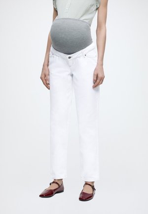 Έγκυος φορά ζώνη εγκυμοσύνης γκρι κάτω από πράσινο καρό πουκάμισο με λευκά τζιν εγκυμοσύνης και κόκκινα παπούτσια Mary Jane.