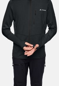 Giacca nera con zip, realizzata in tessuto testurizzato, con fori per il pollice nelle maniche e due tasche con zip. Abbinata a pantaloni scuri e un design minimalista.