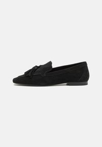 Mocassins de camurça pretos com bico apontado, apresentando um tassel decorativo e uma sola de borracha de baixo perfil. Textura suave em toda a superfície.