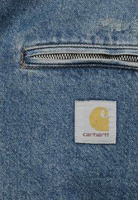 Κοντινό πλάνο μπλε υφάσματος τζιν που δείχνει έναν οριζόντιο μεταλλικό φερμουάρ και μια λευκή ετικέτα της μάρκας Carhartt με κίτρινο λογότυπο.