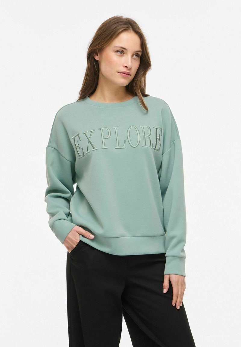 Femme portant un sweat-shirt vert clair avec le texte "EXPLORE", pantalon noir, debout avec une main dans la poche, regardant légèrement vers la droite.