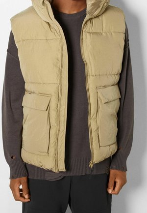Homme portant une veste sans manches matelassée couleur fauve sur un pull à manches longues foncé avec des poignets usés et un pantalon noir.