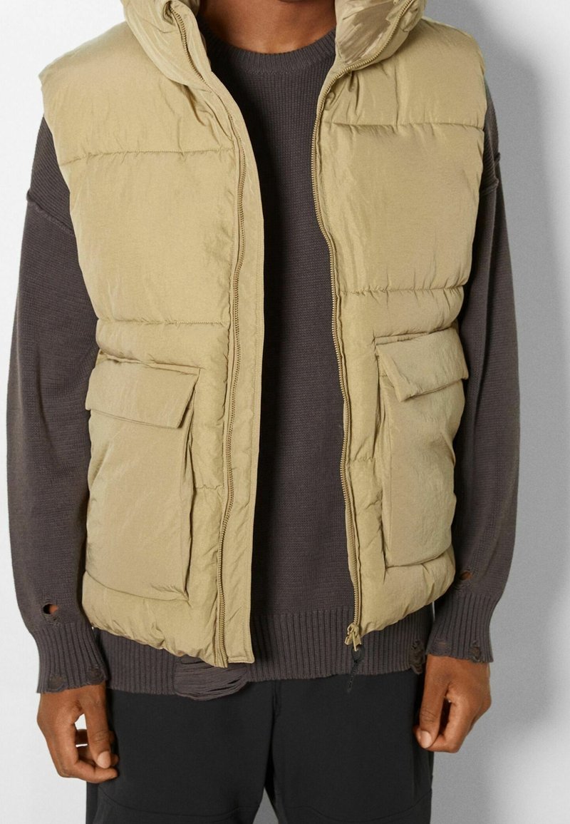 Homme portant une veste sans manches matelassée couleur fauve sur un pull à manches longues foncé avec des poignets usés et un pantalon noir.