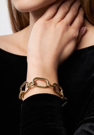 Guld kædearmbånd med sammenkoblede ovale og rektangulære led, båret på et håndled mod en sort fløjlstop. Glat, blank overflade.