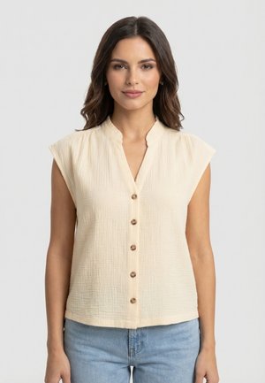 Mujer de cabello oscuro que lleva una blusa sin mangas color crema con botones y jeans azul claro sobre un fondo liso.