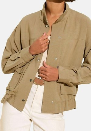 Personne qui ferme les boutons d'une veste fauve à manches longues portée sur un haut beige avec un pantalon blanc et une ceinture marron.