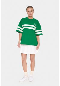 T-shirt verde oversize con strisce orizzontali bianche, maniche corte e collo rotondo, abbinato a una gonna plissettata bianca e scarpe da ginnastica bianche.