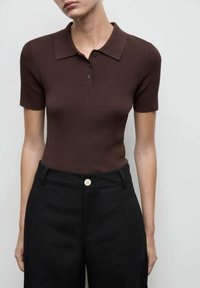 Polo shirt marrone a costine con colletto e maniche corte, abbinato a pantaloni neri a vita alta con un dettaglio di bottone visibile.