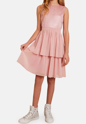 Dziewczyna w bezrękawnej różowej, warstwowej sukience z fakturowanym gorsetem i błyszczącymi wysokimi trampkami, trzymająca brzeg spódnicy.