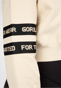 Felpa beige con accenti a strisce nere sulle maniche, caratterizzata da testo bianco. Tessuto morbido con polsini a coste e scollatura arrotondata.