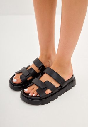 Pieds portant des sandales noires à bouts ouverts avec des semelles épaisses et des brides réglables sur un fond clair.