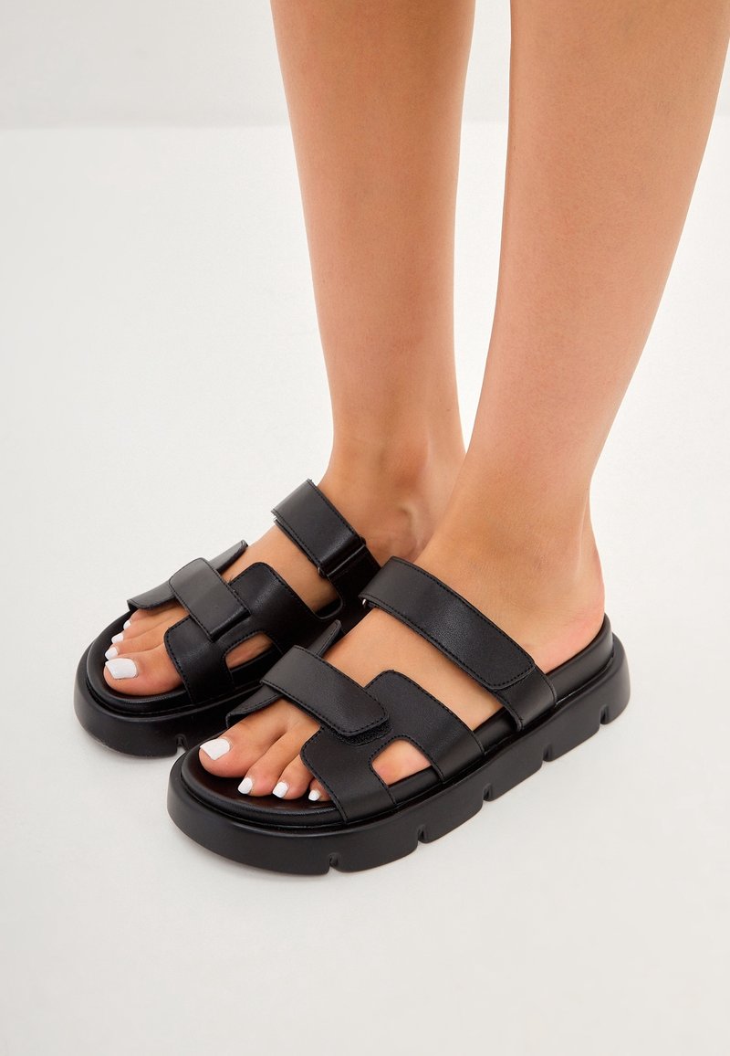Pieds portant des sandales noires à bouts ouverts avec des semelles épaisses et des brides réglables sur un fond clair.