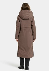 Brun lång parka med huva, tillverkad av slät tyg. Har sidofrall och en åtdragen midja, bärs över mörka byxor och stövlar.