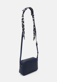 Námořnická obdélníková crossbody taška se zlatým zipem, nastavitelným popruhem a černobílým vzorovaným detailem přivázaným k popruhu.