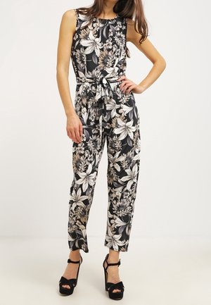 Kvinde iført sort ærmeløs jumpsuit med hvidt og beige blomsterprint og sorte højhælede sandaler mod en ensfarvet baggrund.