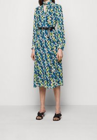 Robe à manches longues ornée d'un imprimé floral multicolore en bleu, vert et blanc, cintrée à la taille par une ceinture noire. Sandales à talons noires.