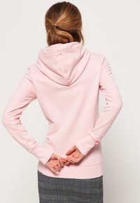 Sudadera rosa claro con capucha ajustable, puños acanalados y un bolsillo de canguro, combinada con una falda a cuadros. Textura de tela suave.