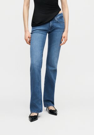 Γυναίκα που φοράει μπλε τζιν παντελόνι bootcut, μαύρο τοπ με πτυχώσεις και μαύρα ψηλοτάκουνα παπούτσια με μυτερή μύτη και φιόγκο, στέκεται σε λευκό φόντο.