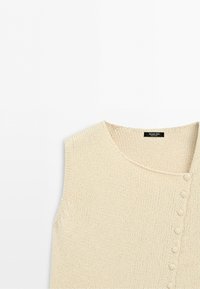 Ärmlös beige stickad topp med rund halsringning och en rad tygklädda knappar längs ena sidan framtill.