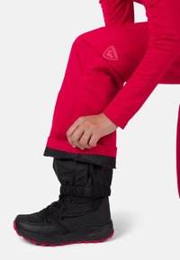 Helle rosa wasserdichte Hose mit einem elastischen Bündchen am Saum, kombiniert mit einem schwarzen Stiefel mit roten Akzenten und Schnürsenkeln, in einer stehenden Position gezeigt.