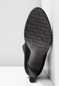 Semelle de chaussure à talon noire avec motif répétitif du logo "Tamaris" et indication de taille 37, chaussure positionnée debout contre un coin blanc.