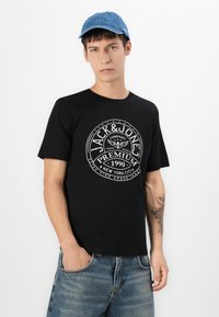 Svart bomulls T-skjorte med en hvit sirkulær logo med tekst. Kombinert med medium vaskede denimshorts og en blå denimcaps.