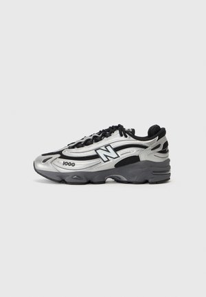 Sølv- og sort New Balance 1100 sneaker med mesh og syntetisk overdel, sorte snørebånd og tyk mørkegrå sål vist fra siden.