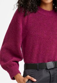 Pull en laine fuchsia à coupe décontractée, poignets côtelés et encolure arrondie, associé à un pantalon noir taille haute et une ceinture texturée.
