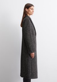 Cappotto grigio con una silhouette lunga, rever a lancia e chiusura a doppio bottone. Presenta spacco laterali e quattro bottoni sui polsini.