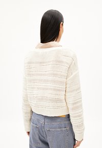 Gekürzter, crèmefarbener Strickpullover mit strukturierten Streifen, gerippten Bündchen und entspannter Passform, getragen über einem khaki- und weißen gestreiften Hemd mit Kragen.