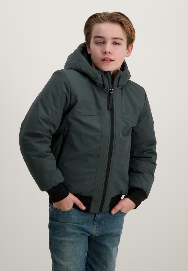 Donkergroene pufferjack met een capuchon, ritssluiting, twee voorkouzijnen en zwarte geribbelde manchetten. Gecombineerd met blauwe jeans.