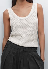 Débardeur en maille beige clair avec un motif en losange, un large décolleté rond et des finitions côtelées, porté avec un pantalon noir à cordon de serrage.