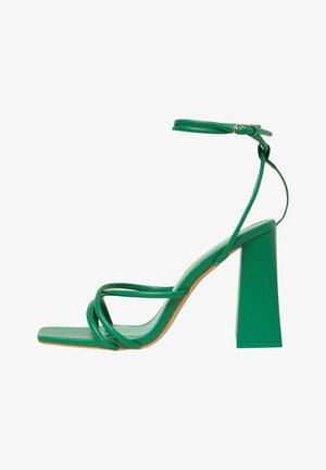 Sandal verde con tacón alto que presenta una punta cuadrada, correas delgadas y una correa para el tobillo con hebilla. El tacón cuadrado es ancho y sólido.