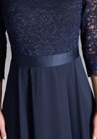 Abito blu navy con corpetto in pizzo, maniche lunghe in pizzo e fascia in satin in vita. Il tessuto plissettato scivola dalla vita, creando una silhouette strutturata.