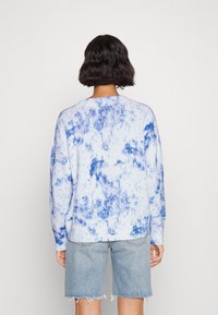 Ljusblått stickat tröja med ett tie-dye-mönster i mörkare blått. Har en ribbad fåll och ärmar, bärs med fransiga denimshorts.