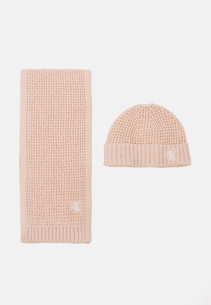 Ensemble châle rose et bonnet assorti. Le châle présente un motif texturé ; le bonnet a un poignet côtelé avec un détail de logo. Les deux sont fabriqués en tissu doux.