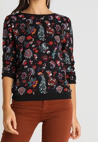 Sudadera negra con vibrantes patrones florales y de pavo real. Presenta mangas largas, puños acanalados y un cuello redondo. Combinada con pantalones marrones.