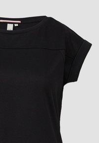 Schwarzes Baumwoll-T-Shirt mit rundem Ausschnitt, kurzen aufgerollten Ärmeln und einem dezenten Nähdetail über der Brust für zusätzliche Textur.