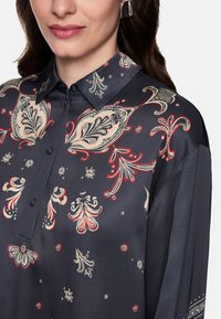 Dark blue blouse with paisley pattern in cream and red. Features a collared neckline and buttons, smooth satin texture, and long sleeves.