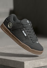 Black synthetic skate shoes with brown rubber soles, featuring embossed logo, white accents, and textured surfaces on the upper.