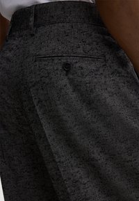 Grijze herringbone broek heeft een gestructureerde stof, een vlakke voorkant en een enkele achterzak met een knoop. De tailleband is op maat gemaakt.