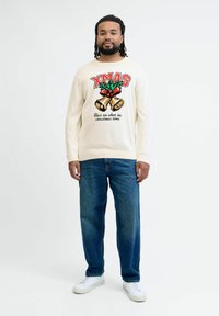 Creme trui met "XMAS" in het rood, feestelijke graphics en tekst, gecombineerd met blauwe denim jeans en witte sneakers. Eenvoudige, casual outfit.