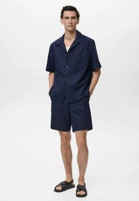 Cămașă scurtă cu mânecă închisă la culoare, pantaloni scurți asortați și sandale negre. Tăietură lejeră, material ușor, textură subtilă și design minimalist.