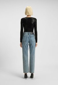 Persoon met kort blond haar die een zwart pailletten top met lange mouwen draagt, lichtblauwe hoge taille jeans met witte spikkels, en zwarte hakken.