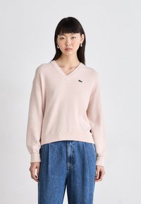 Pull en tricot rose clair avec un col en V, une finition côtelée et des manches longues. Présente un petit logo vert Lacoste sur la poitrine. Assorti à un pantalon en jean bleu.